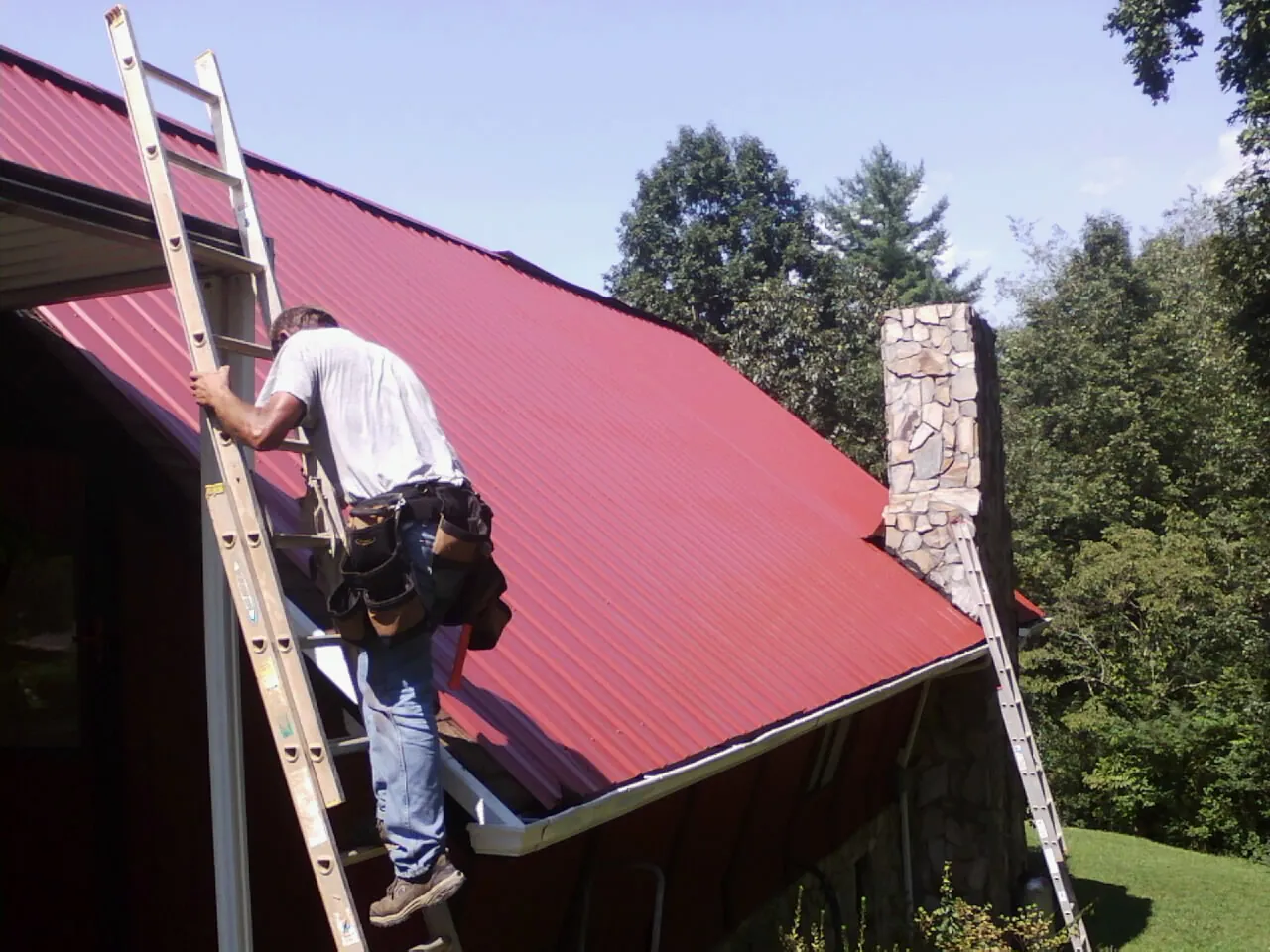Roofer on a ladder beside a red metal roof and stone chimney — Soffit & Fascia Repair by Roofing Brothers in Rehoboth Beach